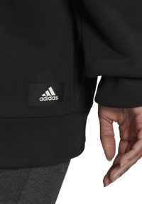 Sudadera negra de Adidas con un parche de logo visible, puños acanalados y un corte relajado, combinada con leggings grises.
