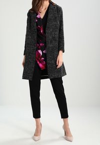 Black and white textured coat with a notched collar, two buttons, and floral wrap dress underneath; paired with black trousers and nude heels.
