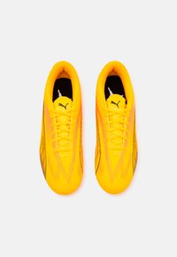 Botas de fútbol amarillas con una textura suave, que presentan franjas diagonales negras y rosas. Diseño con cordones y un logo de Puma en la lengüeta.