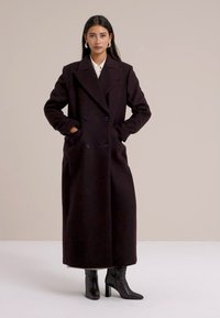 Abrigo largo de doble botonadura de color burdeos oscuro, con grandes bolsillos, solapas en pico y un corte recto. Usado sobre una camisa blanca con cuello y botas negras.