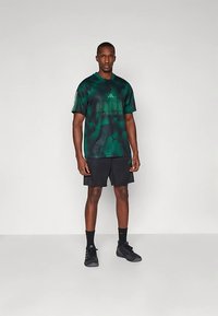 Πράσινη αθλητική φανέλα παραλλαγής με κοντά μανίκια και κείμενο «ADIDAS SPORTSWEAR», συνδυασμένη με μαύρο σορτς και μαύρα αθλητικά παπούτσια.