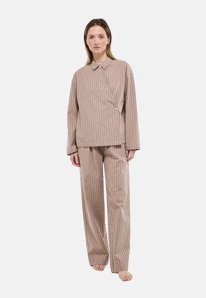 Femme debout pieds nus portant une chemise ample beige à fines rayures et un pantalon large assorti, sur fond blanc.