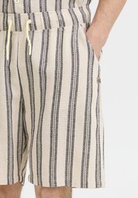 Beige shorts with black stripes, featuring a textured fabric, elastic waistband, and side pockets. Small brand tag on the side.