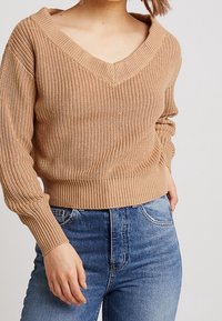 Beige knitted sweater with a wide V-neckline and ribbed cuffs, paired with blue high-waisted denim jeans. Casual fit with textured fabric.