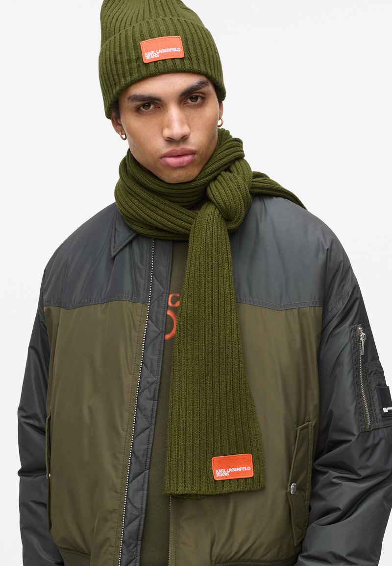 Jovem vestido com um gorro e cachecol de malha verde-azeitona com etiquetas laranja, uma jaqueta bomber num tom verde-azeitona e preta, e brincos de argola dourados.