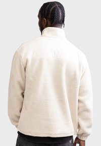 Κρέμ χρώματος φλις τζάκετ με ψηλό γιακά, υφαντό ύφασμα και ελαστικές μανσέτες. Διαθέτει μια αντίθετη λεία πάνελ που διατρέχει τους ώμους.