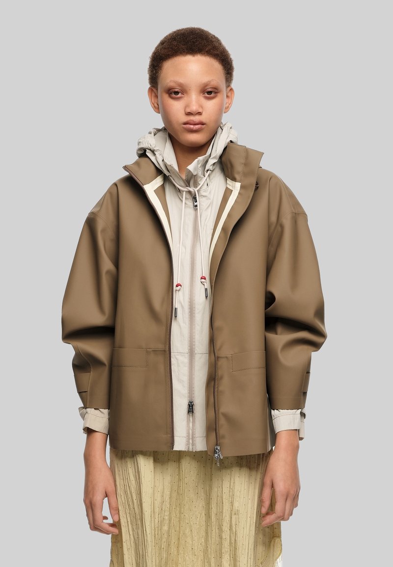 Young person with short hair wearing layered beige and brown jackets over a light yellow pleated skirt against a plain background.