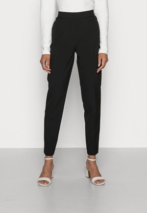 Vrouw die zwarte taps toelopende broek draagt, witte geribbelde top met lange mouwen en witte blokhak sandalen met enkelbandje, staand op een grijze vloer.