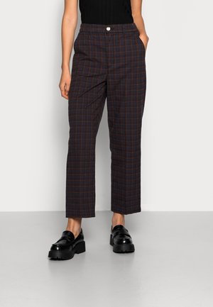 Mujer con pantalones rectos de cuadros oscuros con bolsillos laterales y mocasines negros gruesos, de pie contra un fondo claro y liso.