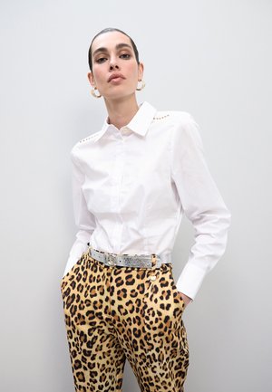 Camisa blanca de botones con detalles en los hombros de tachuelas, combinada con pantalones de estampado de leopardo y un cinturón plateado. Texturas suaves, patrones contrastantes.