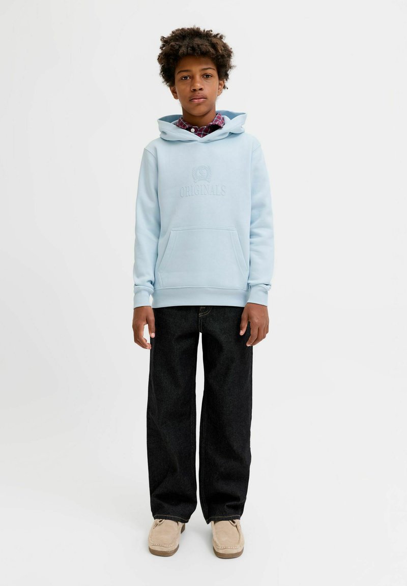 Garçon debout portant un sweat à capuche bleu clair, un jean large foncé, des chaussures beiges et une chemise à carreaux rouge en dessous.