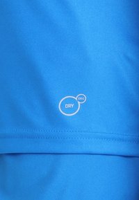 Tejido deportivo azul con una textura suave. Presenta un logo blanco "DRY CELL" impreso en él. Detalle de costura visible en el borde inferior.