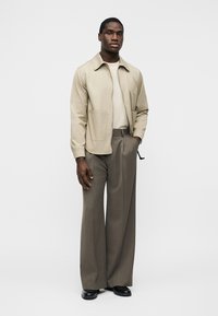 Muž v béžové bundě na zip, krémové košili, širokých hnědých kalhotech a černých botách stojí před jednobarevným světle šedým pozadím.