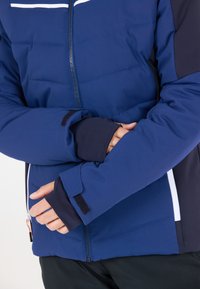 Blå isolerad jacka med hel dragkedja, kontrasterande mörka ärmar och justerbara ärmslut med kardborrstängning. Horisontell vit accentremsa.