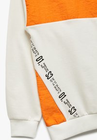 Sweatshirt med en hvid krop og et orange tekstureret øverste panel. Har trykte koordinater og numre i sort langs ærmet.
