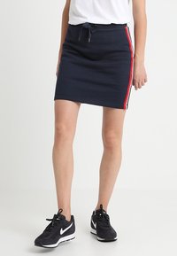 Gonna a maglia blu navy con vita regolabile, strisce laterali rosse e bianche, e design aderente. Abbinata a sneakers nere.