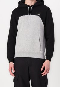 Sudadera con capucha de color negro en la parte superior y gris en el cuerpo, con un bolsillo tipo canguro y cordones. Logo de Lacoste en el pecho. Fabricada en una mezcla de algodón.