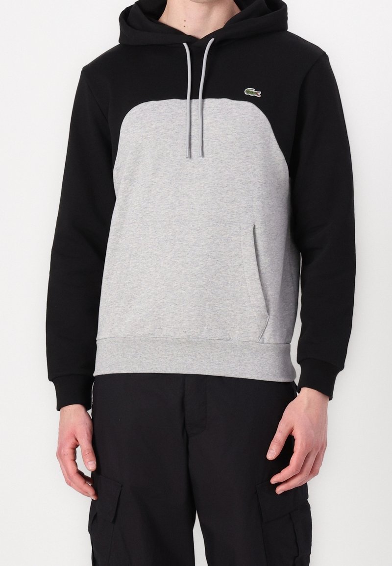 Sudadera con capucha de color negro en la parte superior y gris en el cuerpo, con un bolsillo tipo canguro y cordones. Logo de Lacoste en el pecho. Fabricada en una mezcla de algodón.