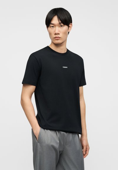 Νέος άνδρας με ίσια μαύρα μαλλιά φοράει μαύρο T-shirt με κοντά μανίκια και το λογότυπο "ICEBERG" καθώς και φαρδιά γκρι παντελόνια, με τα χέρια στις τσέπες.