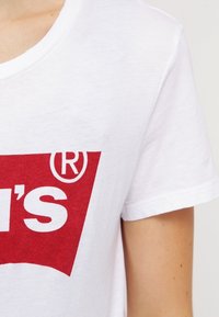 Bílé bavlněné tričko s velkým červeným logem a textem "LEVI'S" s křivým akcentem. Tkanina má hladký povrch a krátké rukávy.