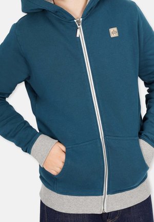 Haut à zip teal avec un ourlet et des poignets côtelés gris, présentant un patch logo sur la poitrine et une capuche doublée. Le matériau semble doux.