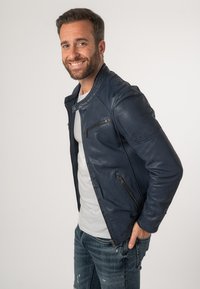 Giacca di pelle blu navy con un design aderente, caratterizzata da una tasca con zip e colletto alto; abbinata a una maglietta grigia e jeans strappati.
