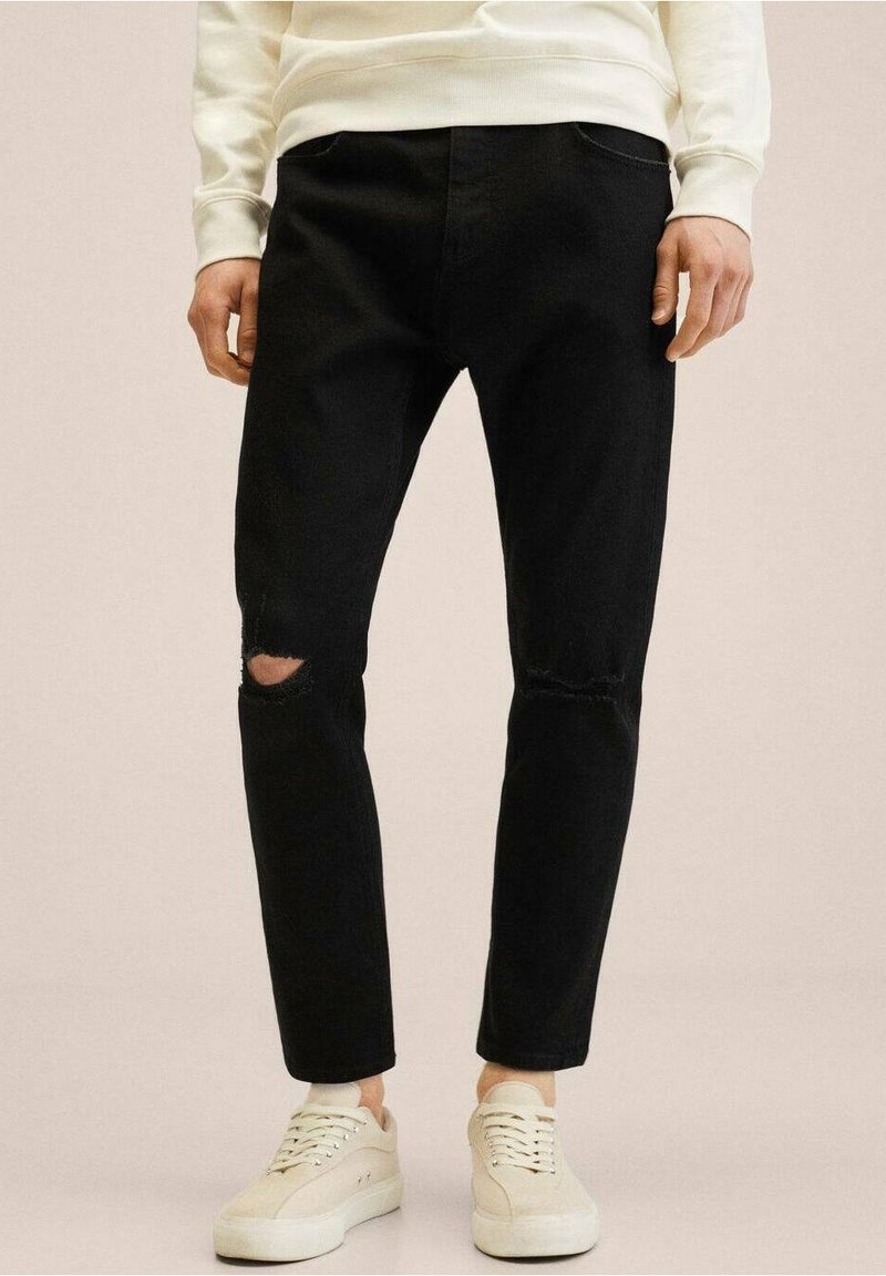 Vaqueros de mezclilla negros con desgarros visibles en las rodillas, ajuste slim y diseño clásico de 5 bolsillos, combinados con zapatillas crema sobre un fondo neutro.