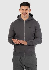 Haut de sweatshirt zippé gris avec capuche, poignets et ourlet côtelés. Présente un petit logo noir sur la poitrine. Tissu chaud et doux, design minimal.