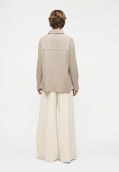 Άτομο με κοντά σγουρά μαλλιά φοράει μπεζ πουκάμισο με κουμπιά και κρεμ παντελόνι φαρδύ, στέκεται μπροστά σε λευκό, απλό φόντο, με την πλάτη γυρισμένη.