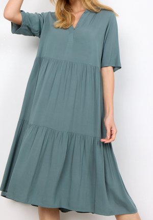 Femme portant une robe midi ample, à étages, bleu sarcelle, avec manches courtes et décolleté en V, debout sur un fond blanc uni.