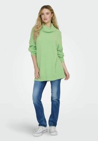 Hele rohelise oversize-sviiter, millel on kaelaosa, tekstureeritud kangas ja soonikkoes äär, koos siniste teksade ja valgete tossudega.