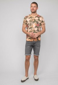 Chemise à manches courtes de couleur pêche avec un motif floral vert et rose, associée à un short en jean gris roulé et des chaussures à enfiler beige.