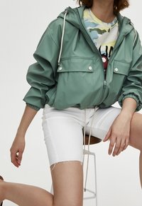 Chaqueta ligera con capucha en verde menta, con bolsillos frontales, puños elásticos y cierre de cremallera, combinada con pantalones cortos blancos sobre un taburete.