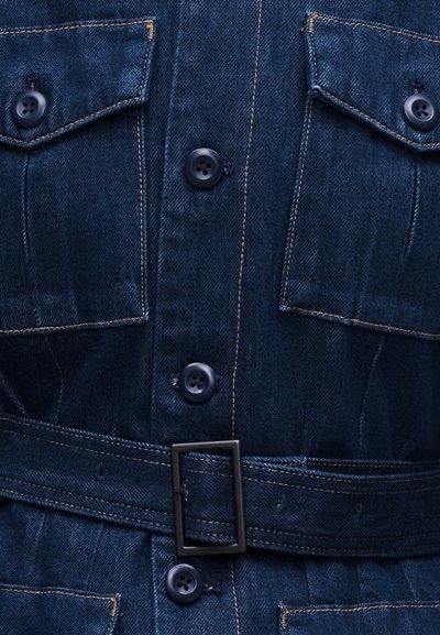 Σκούρο μπλε τζιν ενδυμα με μαύρα κουμπιά, δύο τσέπες στο στήθος με καλύμματα, και μια ταιριαστή ζώνη με ορθογώνια μαύρη αγκράφα.
