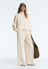 Donna che indossa una giacca beige con zip, pantaloni larghi abbinati, cappellino grigio, scarpe beige, con una grande borsa marrone scuro, su uno sfondo neutro.