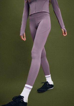 Donna che indossa leggings color malva e una giacca con zip, abbinata a calzini bianchi e sneakers nere, su uno sfondo verde oliva.