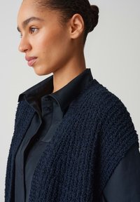 Gilet blu impermeabile realizzato in tessuto morbido, indossato sopra una camicia nera con colletto. Il gilet presenta una vestibilità ampia con un design a maglia grossa.