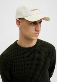 Een lichtbeige baseballcap met "Originals" geborduurd in koper. Draagt een donkergroene, gestructureerde breisweater.