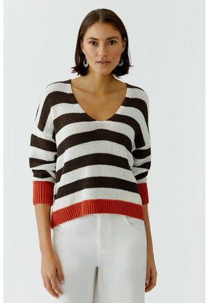 Vrouw draagt een bruin-wit horizontaal gestreepte trui met oranje manchetten en zoom, gecombineerd met een witte broek, staand tegen een effen achtergrond.