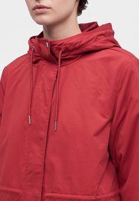 Personne portant une veste à capuche rouge avec cordons et fermeture à pression, montrée du cou jusqu'au milieu du torse.
