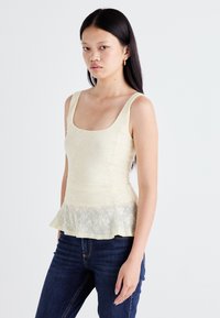 Een model in een lichtgele kanttop en donkere jeans poseert tegen een neutrale achtergrond, waarbij het peplumontwerp en de getailleerde pasvorm van de top worden getoond.