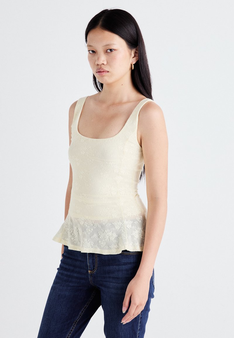 Een model in een lichtgele kanttop en donkere jeans poseert tegen een neutrale achtergrond, waarbij het peplumontwerp en de getailleerde pasvorm van de top worden getoond.