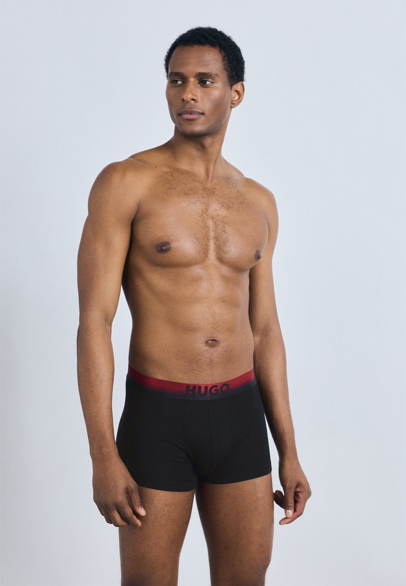 Boxer noirs avec une ceinture élastique à dégradé rouge arborant la marque "HUGO". Le modèle pose avec une expression neutre et une silhouette définie.