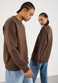 To personer med flettehår som har på seg brune sweatshirts og lyseblå jeans står mot en enkel hvit bakgrunn.