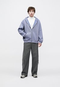 Sweat-shirt com capuz e fecho éclair em roxo claro, com capuz, grandes bolsos, combinado com calças cargo cinzentas com fechos éclair laterais e ténis cinzentos.