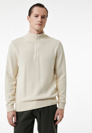 Junger Mann, der einen cremefarbenen Halb-Reißverschluss-Pullover und dunkle Hosen trägt, steht vor einem schlichten weißen Hintergrund.