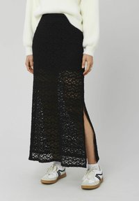 Jupe longue noire en maille avec un motif en dentelle semi-transparent et des fentes latérales. Portée avec des baskets blanches ornées d'accents noirs.