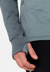Langarmshirt aus meliertem Teal-Stoff, mit Daumenlöchern, strukturiertem Muster und einer enganliegenden Passform. Tragen mit schwarzen Sportleggings.