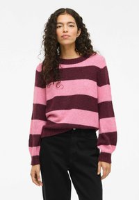 Lila und rosa gestreifter Strickpullover mit Rundhalsausschnitt, geripptem Saum und Bündchen, kombiniert mit schwarzen Hosen, die eine weiche Textur zeigen.