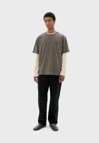 Graues Baumwoll-T-Shirt mit Rundhalsausschnitt, über einem cremefarbenen Langarmshirt getragen. Schwarze, locker sitzende Hose und graue Slipper vervollständigen das Outfit.